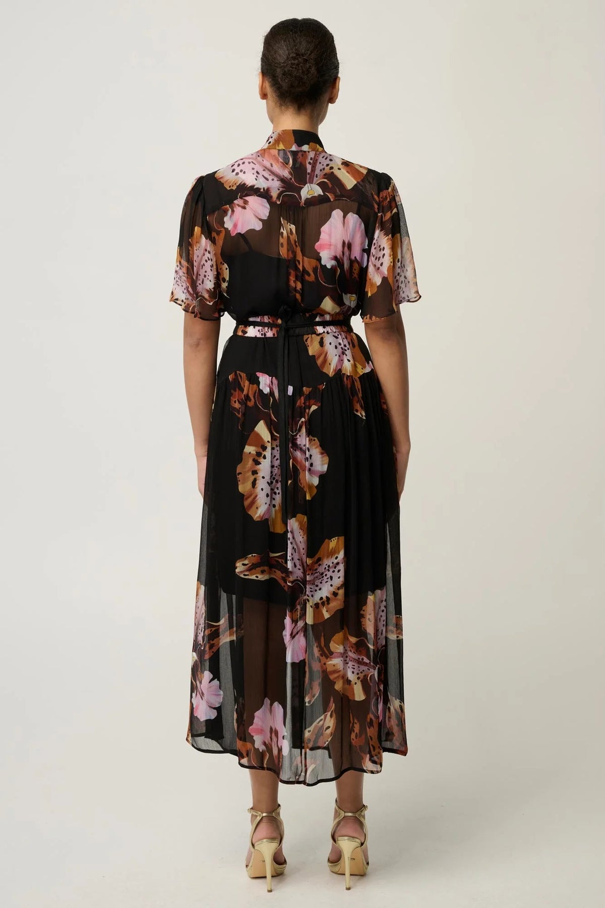 Woman wearing a black floral floaty chiffon dress with tie waist and short sleeves and gold sandals against a white background.