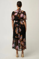 Woman wearing a black floral floaty chiffon dress with tie waist and short sleeves and gold sandals against a white background.