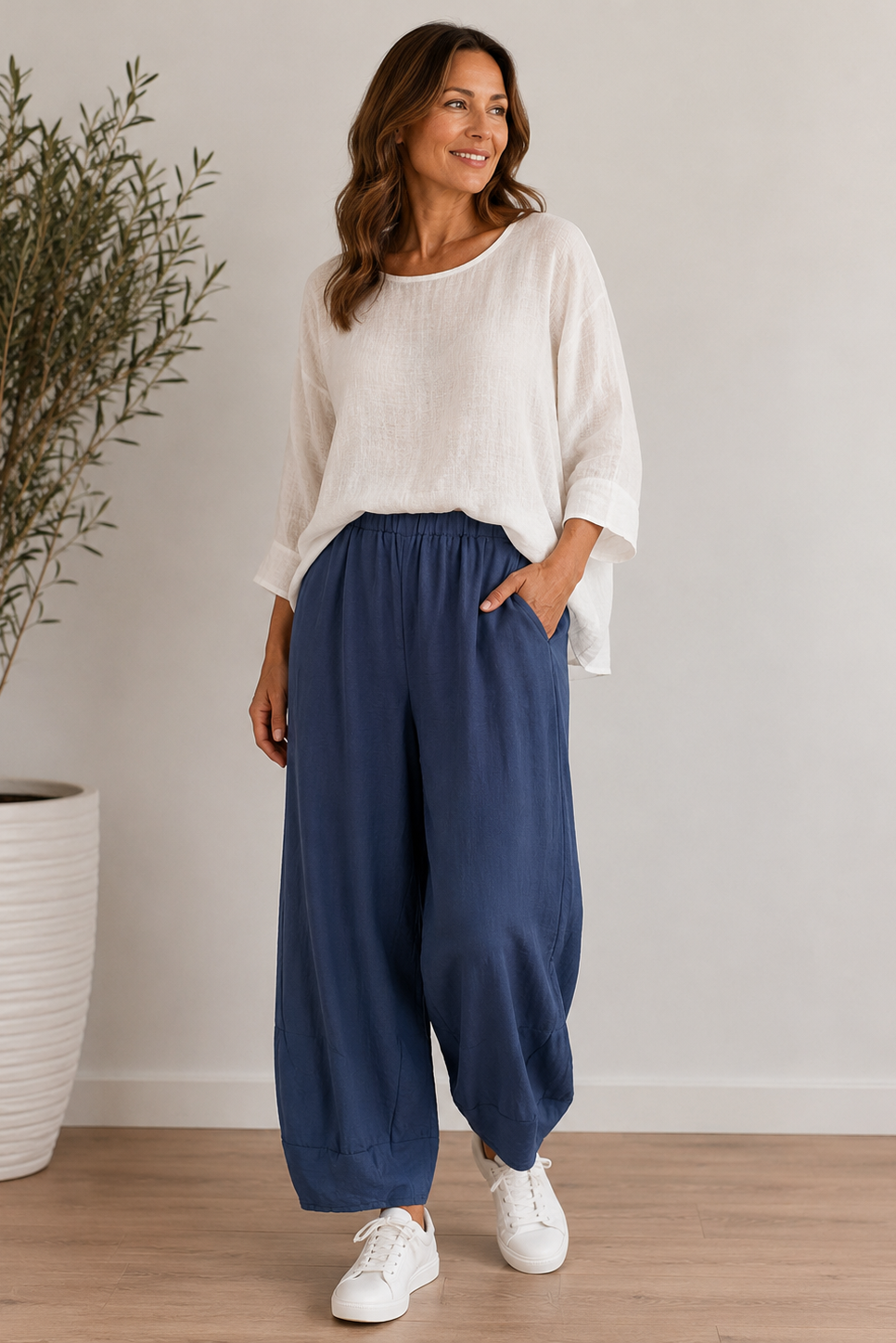 A woman modeling a white long-sleeve linen top and navy blue wide-leg pants, standing in a bright room with a wooden floor and a potted plant in the background.