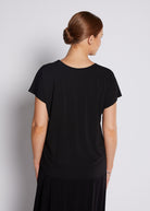 A woman viewed from the back wearing a black short-sleeve top and a black skirt against a light gray studio background.