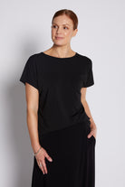 A woman modeling a black short-sleeve boat-neck top and black skirt against a light gray studio background.