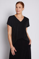 A woman modeling a black short-sleeve V-neck top with an asymmetrical draped hem, paired with a black skirt, against a light gray studio background.