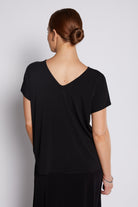 A woman viewed from the back wearing a black short-sleeve V-neck top and black skirt against a light gray studio background.