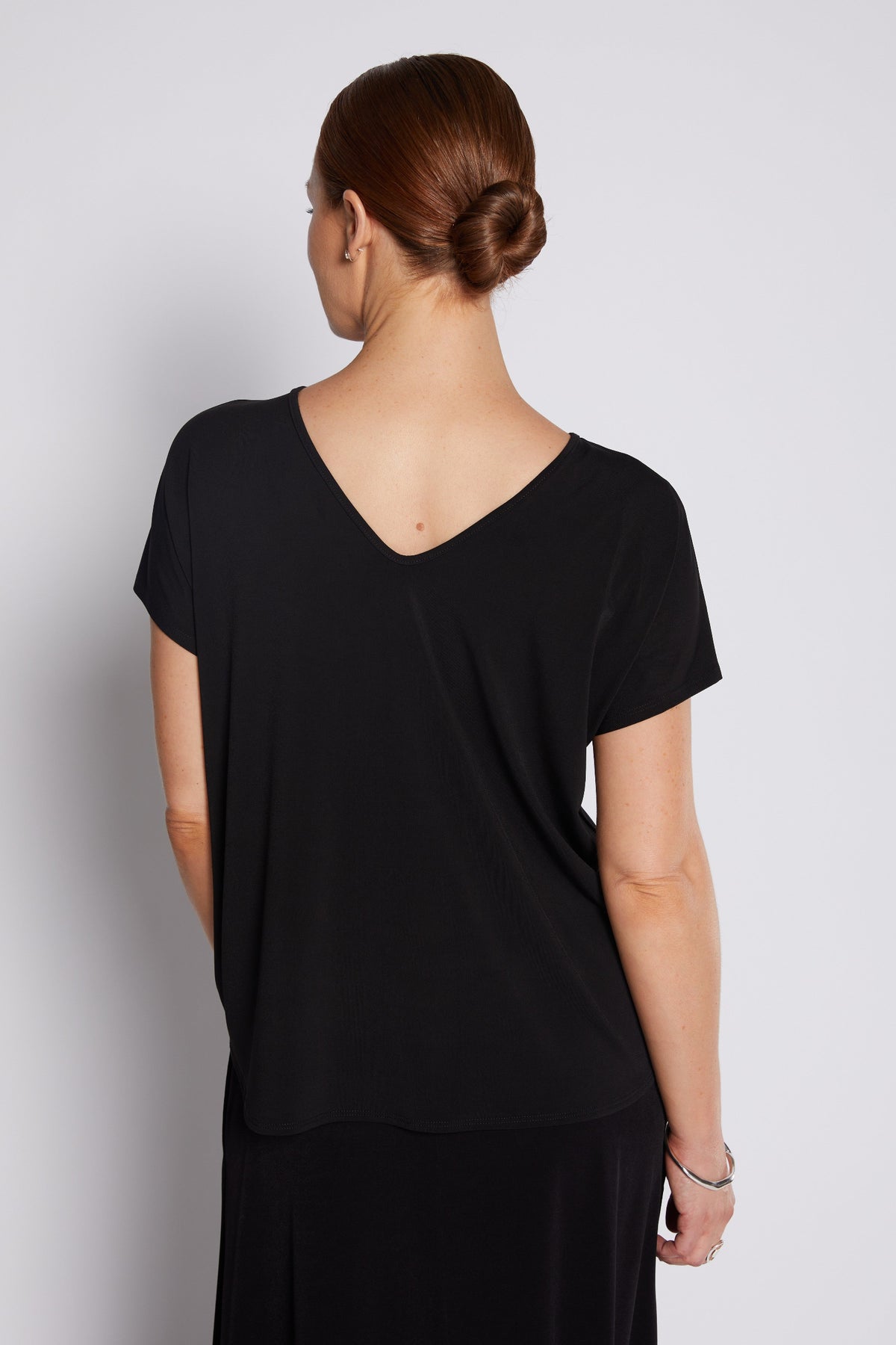 A woman viewed from the back wearing a black short-sleeve V-neck top and black skirt against a light gray studio background.