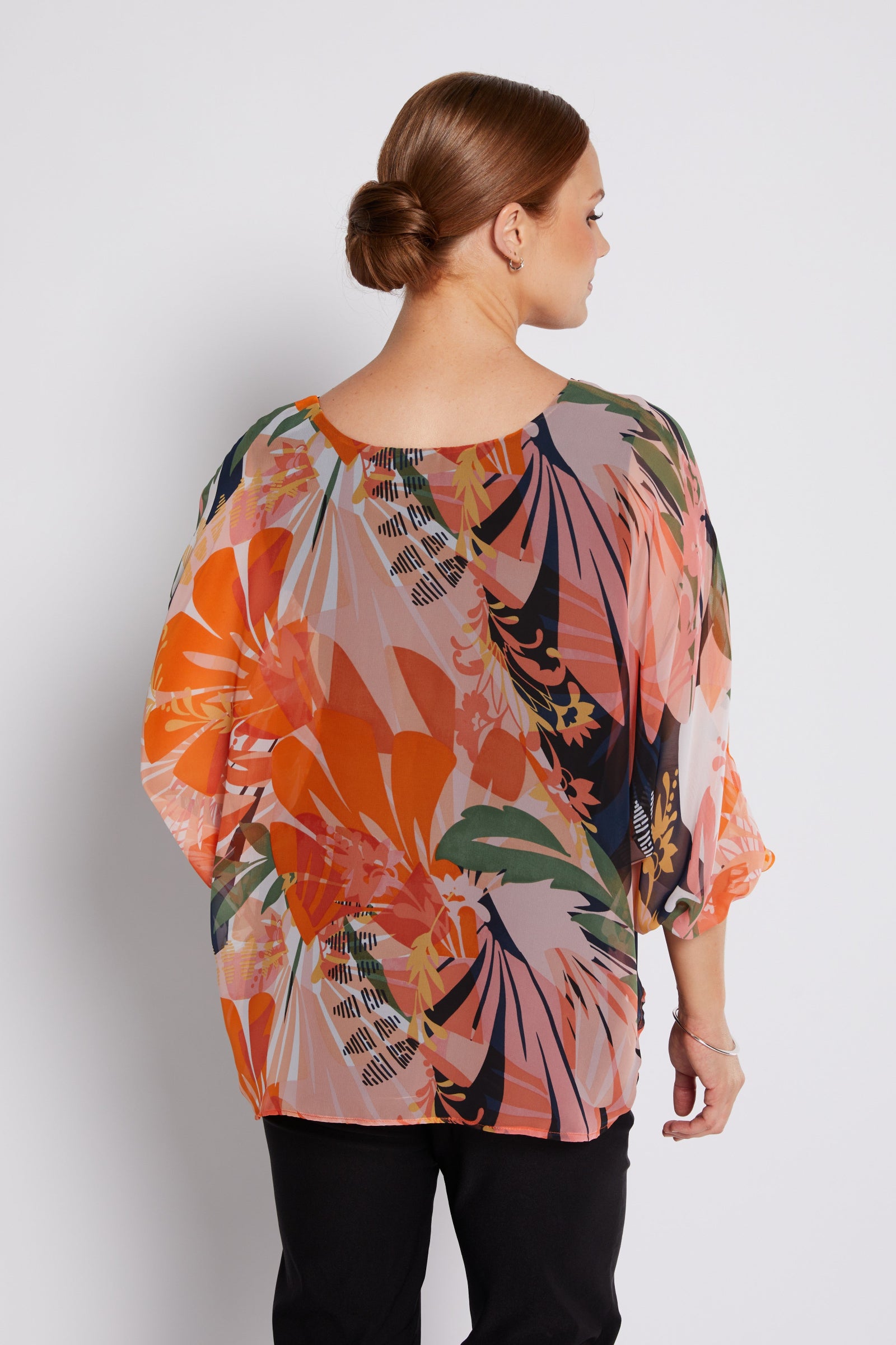 A woman shown from the back wearing a loose, short-sleeved blouse with a vibrant tropical floral print in orange, pink, green, and navy on a light beige base, paired with black pants against a light gray studio background.