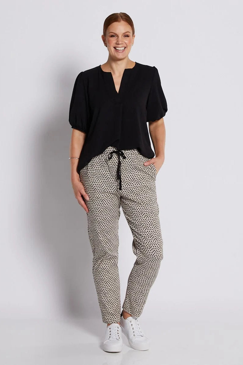 A woman modeling a black short-sleeve top with a split neckline and patterned jogger pants featuring a black and beige geometric print, paired with white sneakers, against a light gray studio background.