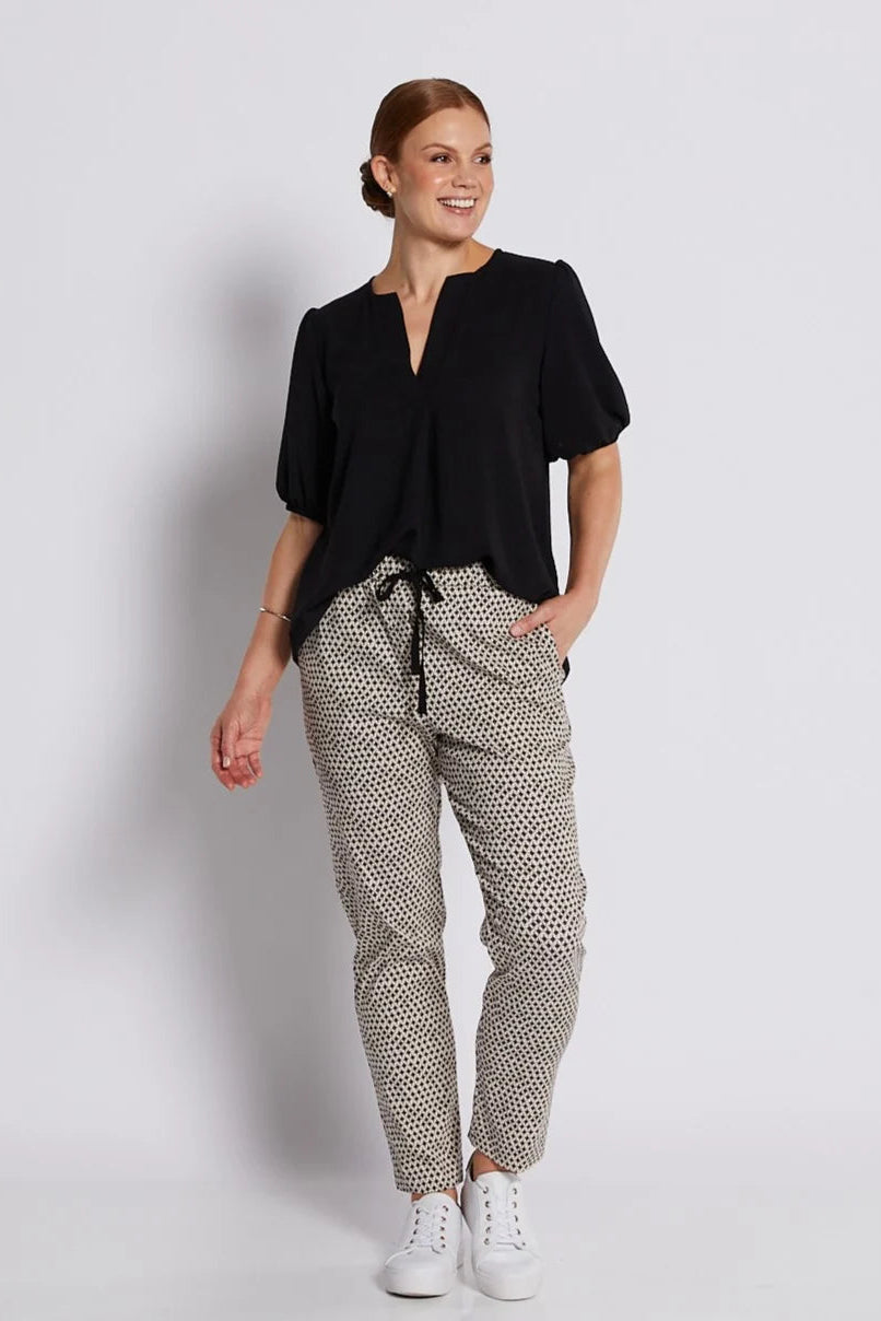 A woman modeling a black short-sleeve blouse with a split neckline and patterned drawstring trousers featuring a black and beige geometric print, paired with white sneakers, against a light gray studio background.
