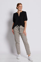 A woman modeling a black short-sleeve blouse and beige-and-black patterned trousers with white sneakers, standing against a light gray studio background.
