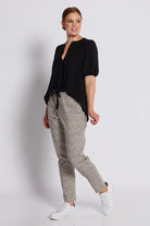 A woman modeling a black short-sleeve blouse and beige-and-black patterned trousers with white sneakers, standing against a light gray studio background.
