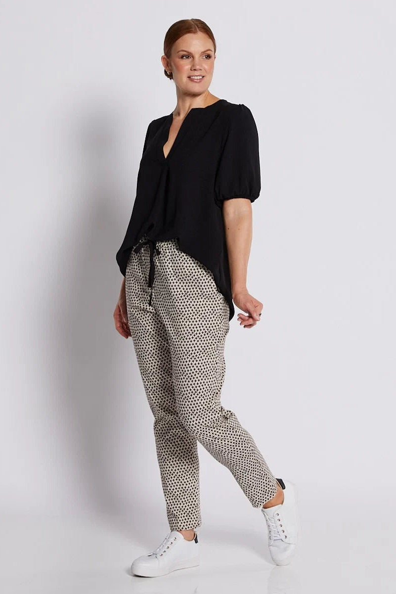 A woman modeling a black short-sleeve blouse and beige-and-black patterned trousers with white sneakers, standing against a light gray studio background.