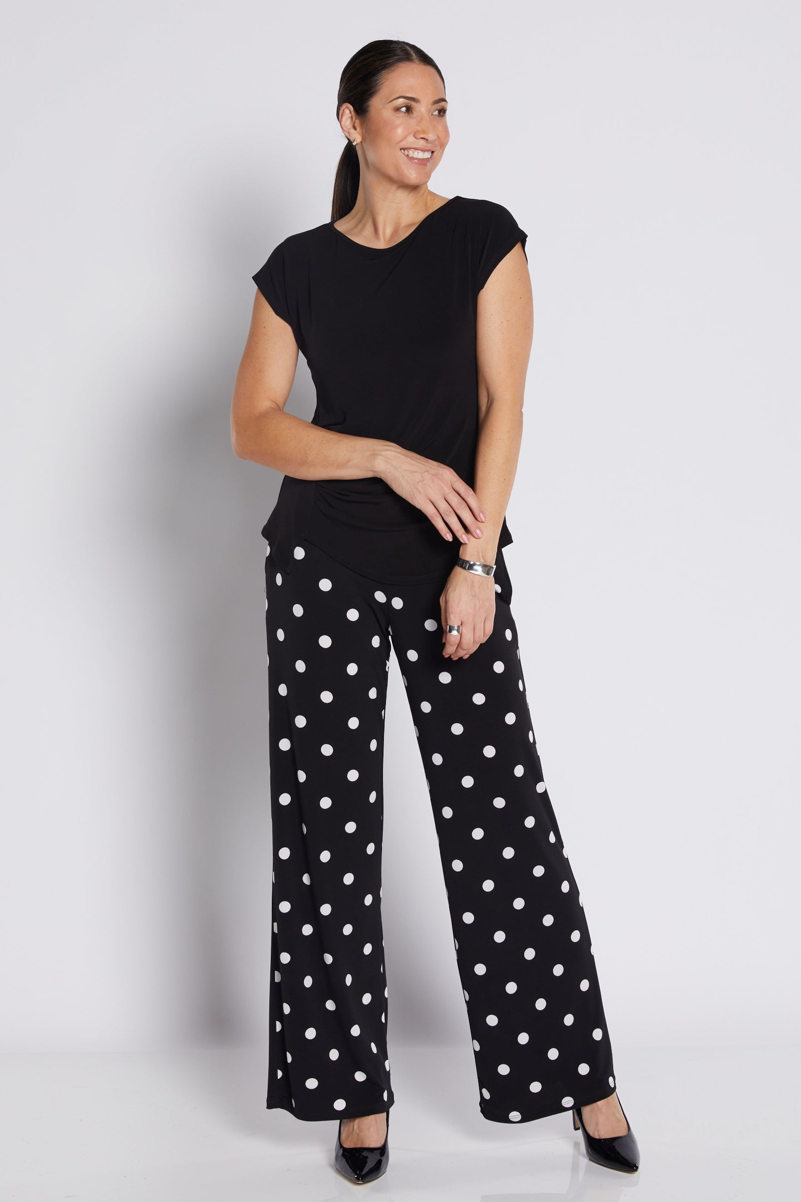 A woman modeling a black short-sleeve top and black wide-leg pants with white polka dots, standing against a light gray studio background.