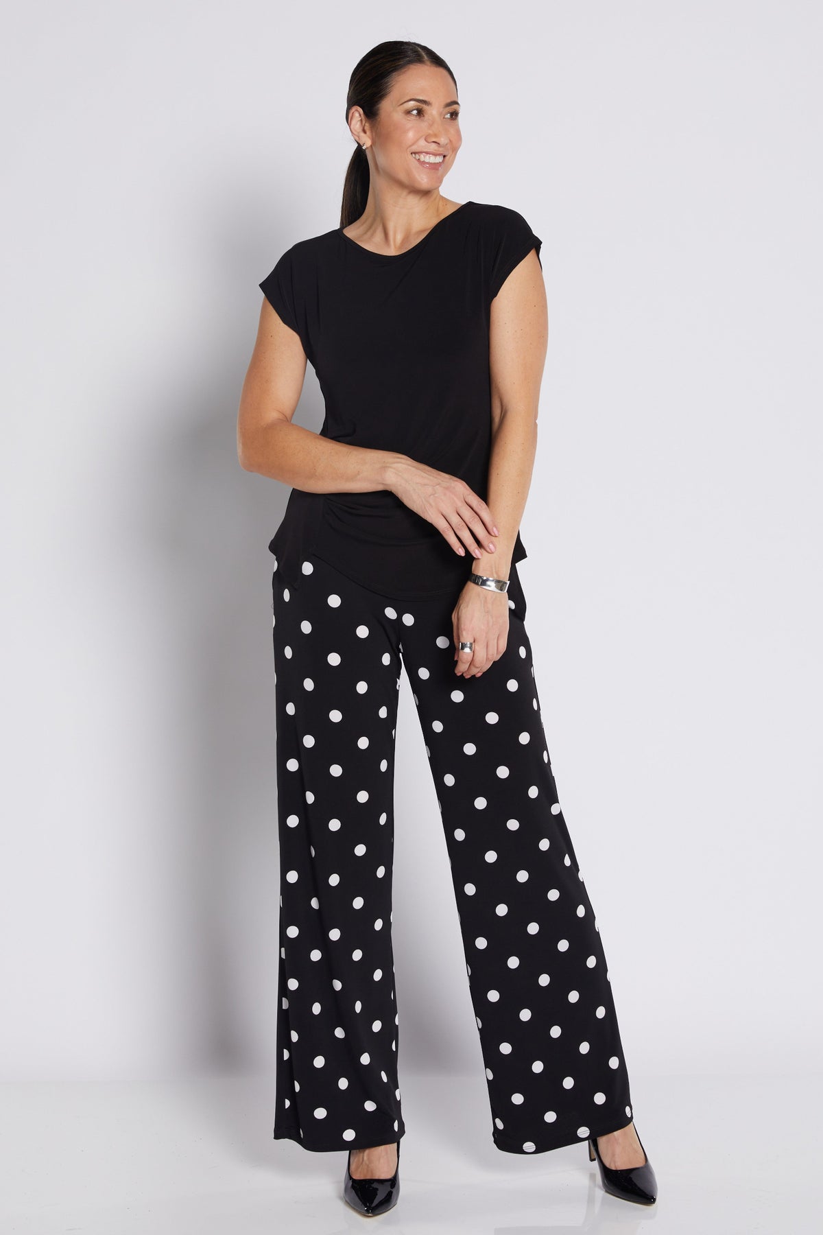 A woman modeling a black short-sleeve top and black wide-leg pants with white polka dots, standing against a light gray studio background.