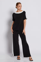 A woman modeling a black two-piece outfit consisting of a short-sleeve top with white contrast trim at the neckline and sleeve cuffs, and matching black wide-leg pants with white side panels, against a light gray studio background.