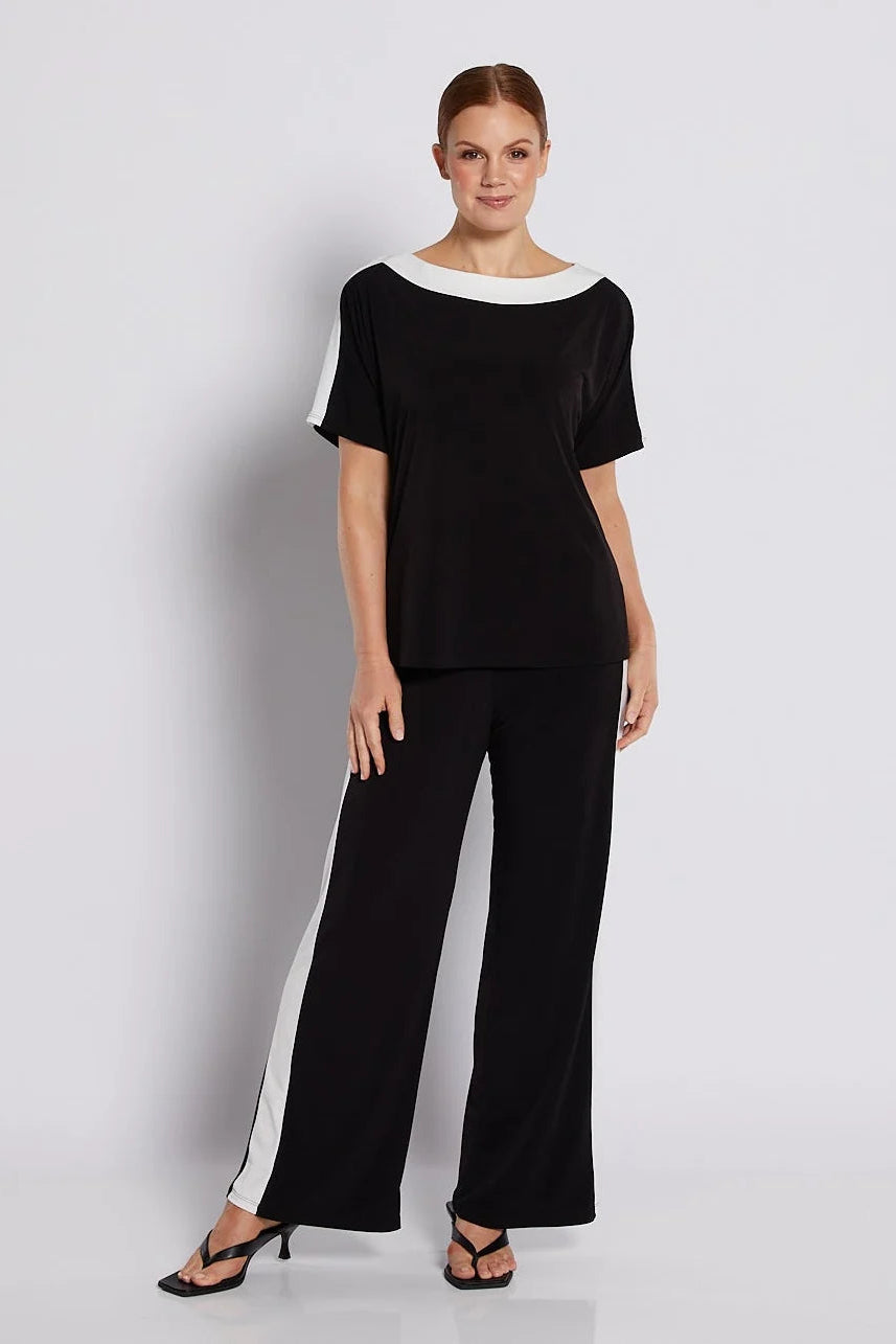 A woman modeling a black two-piece set consisting of a short-sleeve top with a white boat neckline and white side panels, paired with matching black wide-leg pants featuring white side panels, against a light gray studio background.