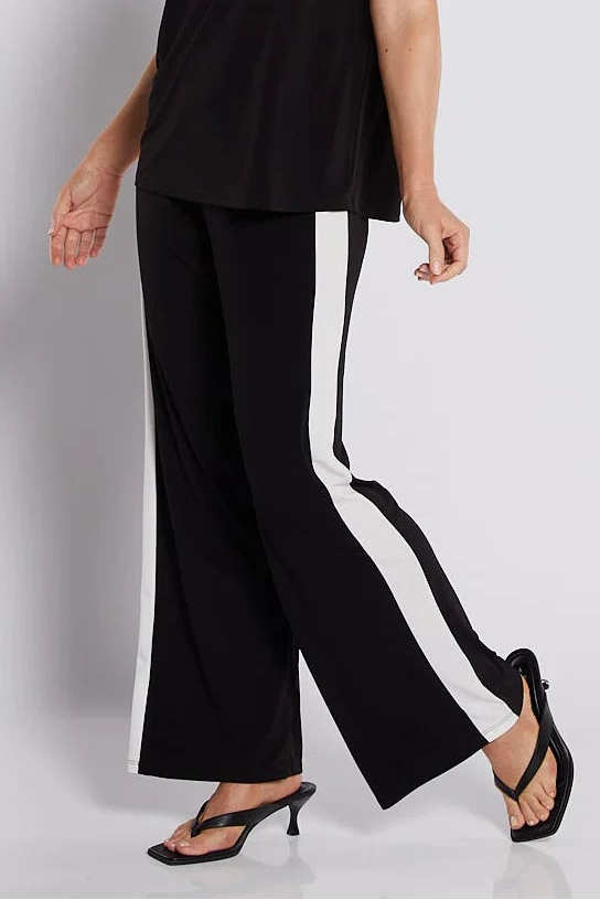 A woman modeling a black two-piece set consisting of a short-sleeve top with white shoulder stripes and matching wide-leg trousers with white side stripes, paired with black strappy heels, against a light gray studio background.