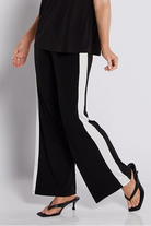 A woman modeling a black two-piece set consisting of a short-sleeve top with white shoulder stripes and matching wide-leg trousers with white side stripes, paired with black strappy heels, against a light gray studio background.