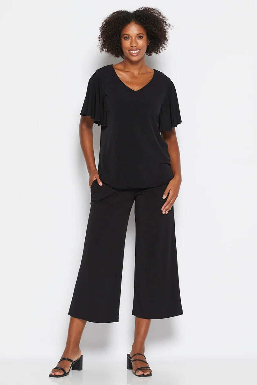 A woman modeling a black two-piece outfit consisting of a short-sleeve V-neck top with ruffled sleeves and matching wide-leg cropped pants, paired with black heeled sandals against a light gray studio background.