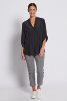 A woman modeling a black long-sleeve button-up shirt with pleated detailing and black-and-white gingham cropped pants, paired with white sneakers, against a light gray studio background.