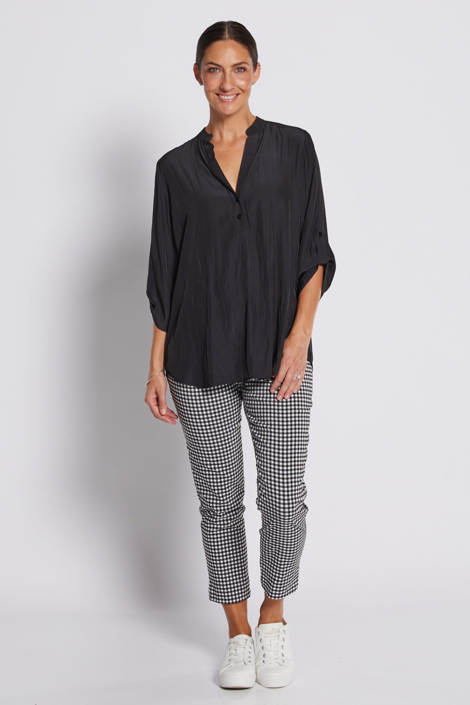 A woman modeling a black long-sleeve button-up shirt with pleated detailing and black-and-white gingham cropped pants, paired with white sneakers, against a light gray studio background.