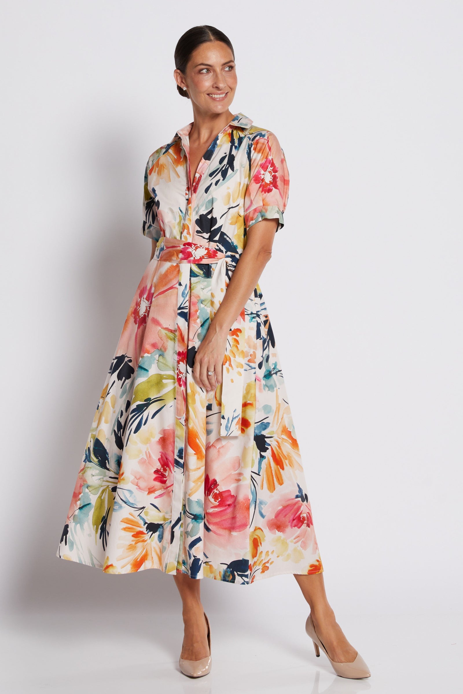 A woman modeling a short-sleeved midi dress with a colorful floral print, featuring a tie waist belt and a V-neckline, set against a plain light gray studio background.