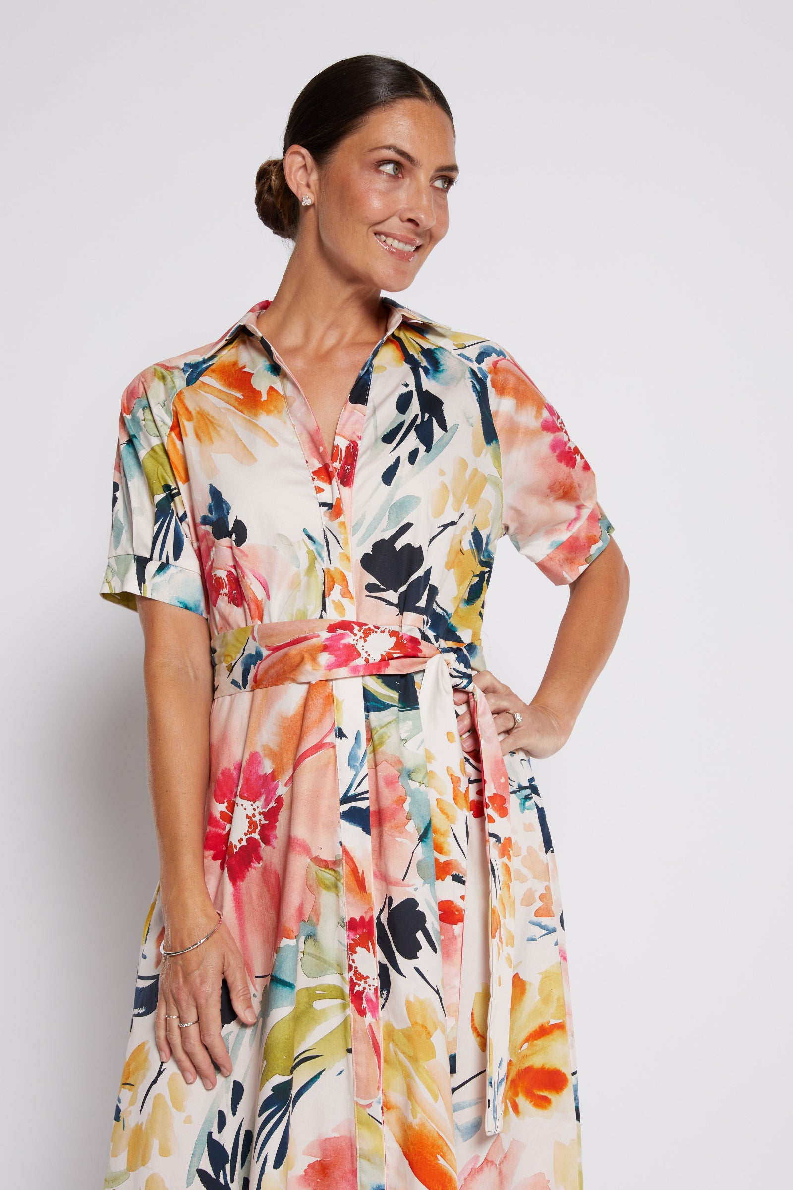 A woman modeling a short-sleeve collared dress with a colorful floral print, featuring a self-tie waist belt, set against a plain light gray studio background.