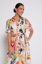 A woman modeling a short-sleeve collared dress with a colorful floral print, featuring a self-tie waist belt, set against a plain light gray studio background.