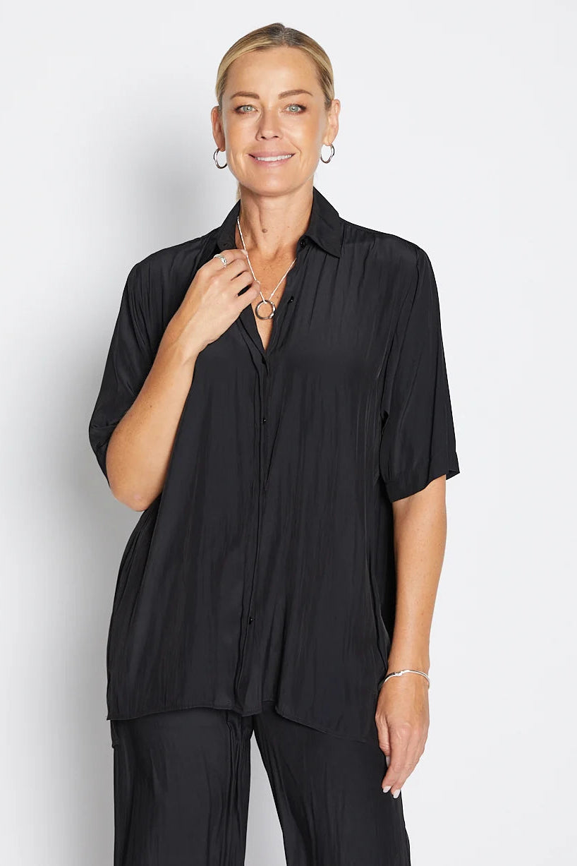 A woman modeling a black short-sleeve button-up shirt with matching black trousers, photographed against a light gray studio background.