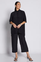 A woman modeling a black two-piece outfit consisting of a short-sleeve, high-neck, pleated blouse and matching cropped pants, paired with black sandals against a light gray studio background.