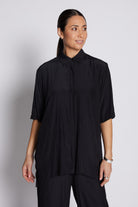 A woman modeling a black short-sleeve button-up shirt with a high collar and subtle vertical pleating, paired with black trousers, against a light gray studio background.