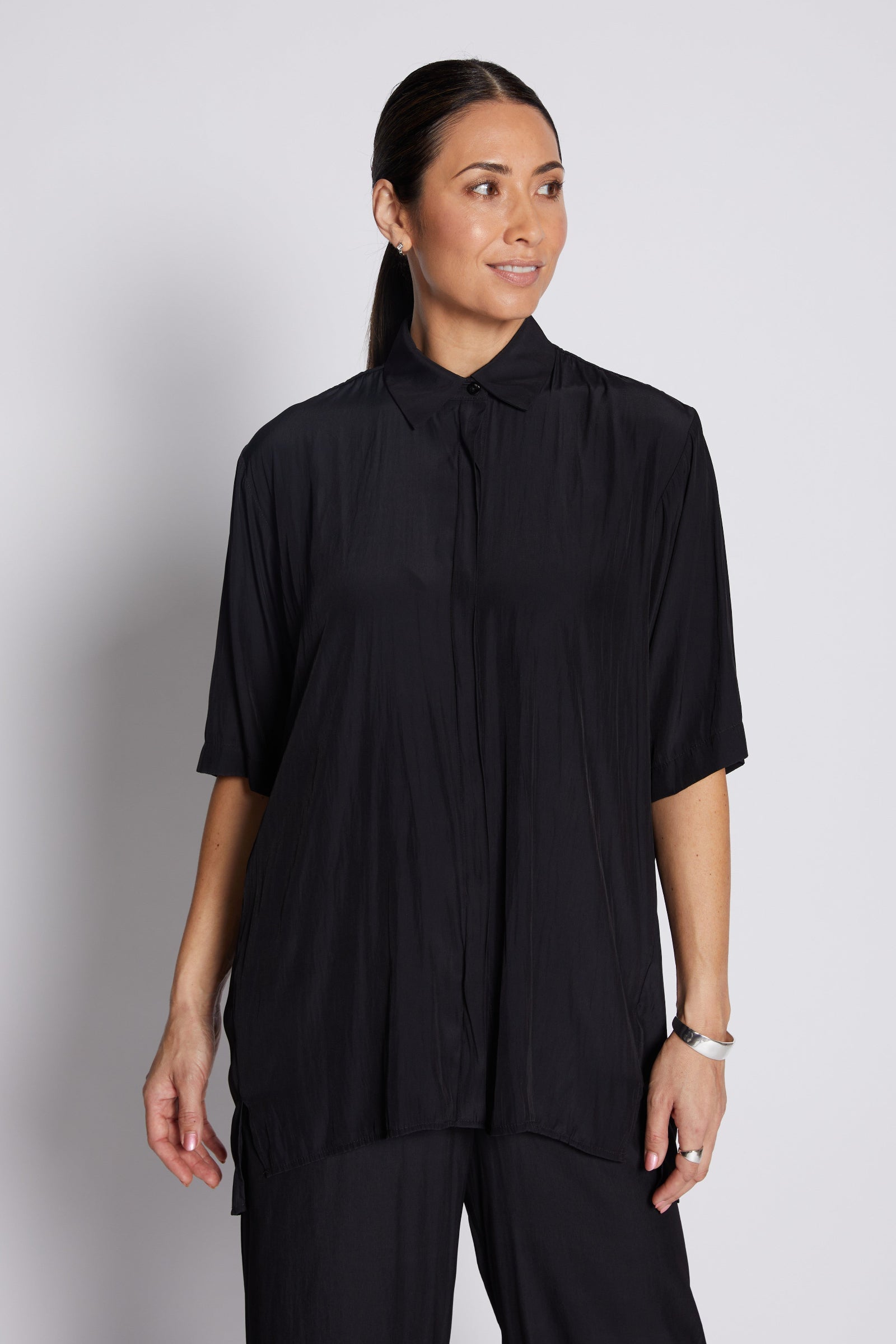 A woman modeling a black short-sleeve button-up shirt with a high collar and subtle vertical pleating, paired with black trousers, against a light gray studio background.