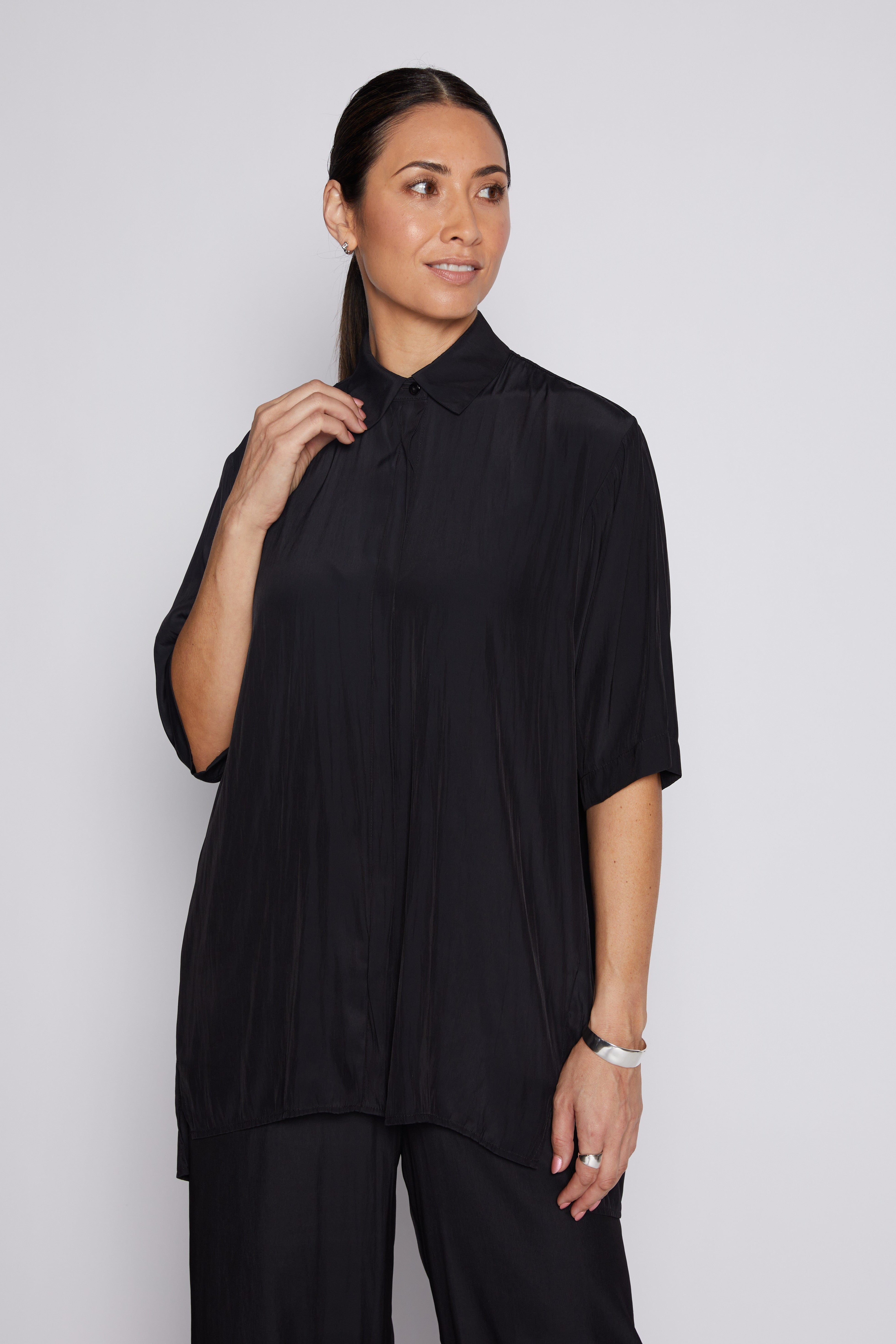 A woman modeling a black short-sleeve pleated button-up shirt with a collar, paired with black trousers, against a light gray studio background.