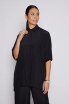 A woman modeling a black short-sleeve pleated button-up shirt with a collar, paired with black trousers, against a light gray studio background.