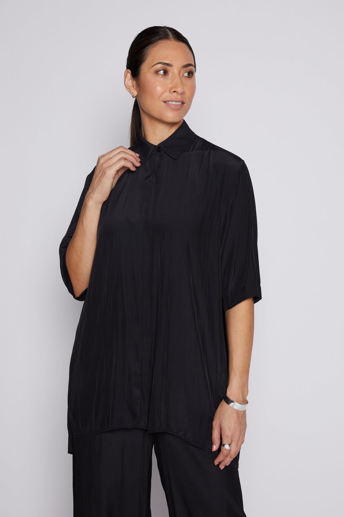 A woman modeling a black short-sleeve pleated button-up shirt with a collar, paired with black trousers, against a light gray studio background.
