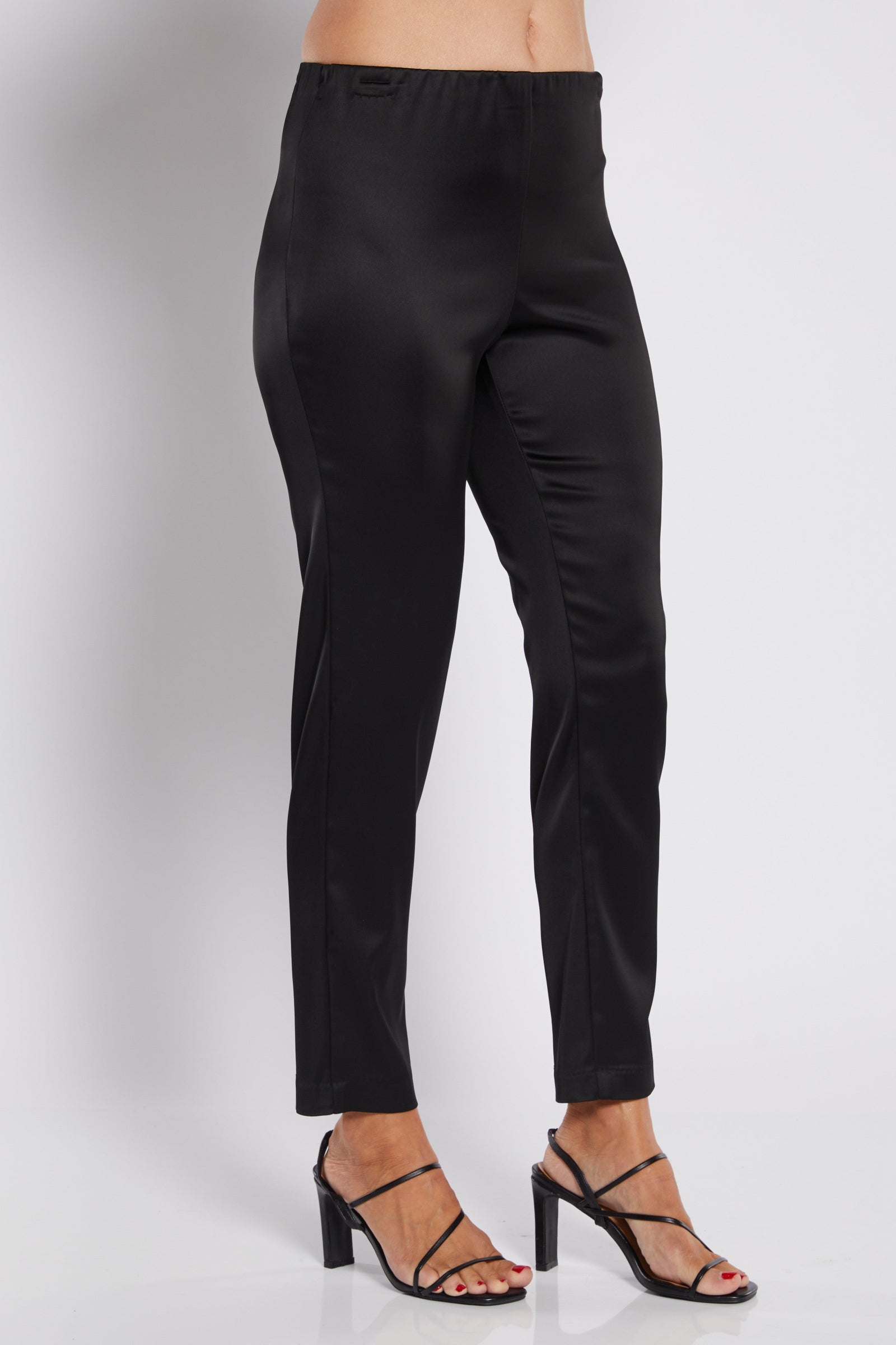 Black slim-fit ankle-length pants shown on a model from the waist down, paired with black strappy high-heeled sandals against a light gray studio background.