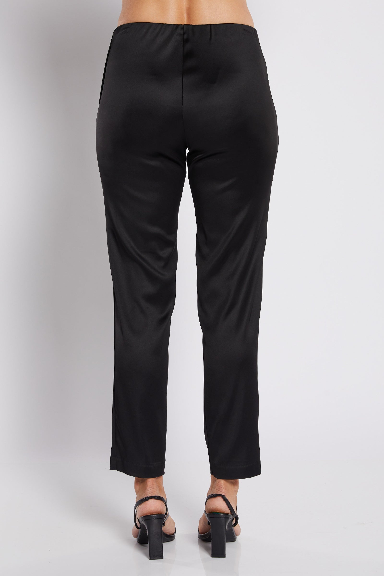 Rear view of a model wearing black satin trousers and black block-heel sandals against a light gray studio background.