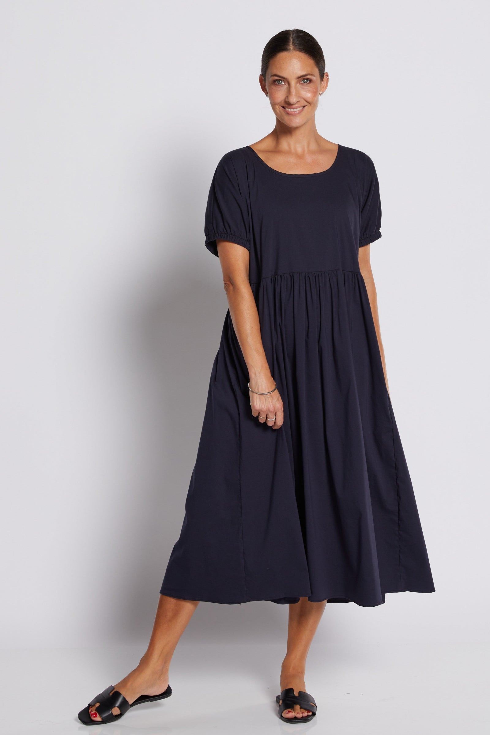 A woman modeling a short-sleeve, scoop-neck midi dress in dark navy blue, paired with black slide sandals, against a light gray studio background.