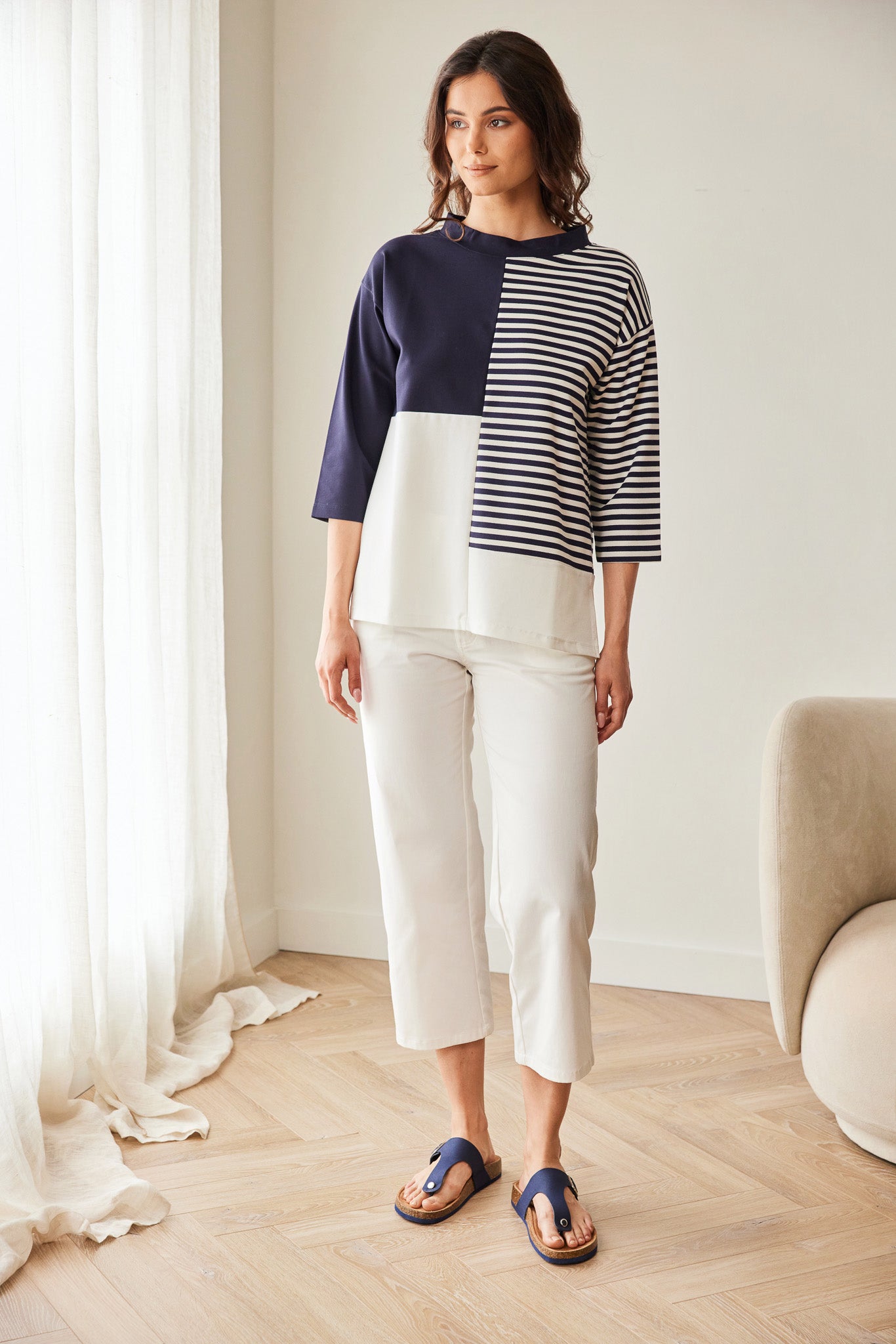 Woman wearing a color-blocked sweater and white pants in a room with light wood flooring and a white wall.