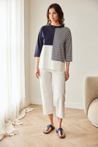 Woman wearing a color-blocked sweater and white pants in a room with light wood flooring and a white wall.