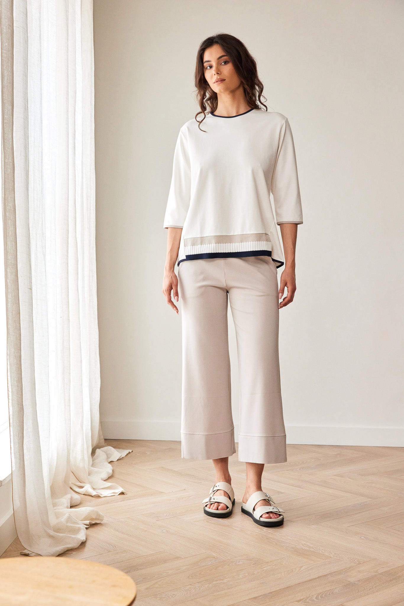 A woman stands in a bright room wearing a cream-colored knit top and matching wide-leg cropped pants, paired with white slide sandals.