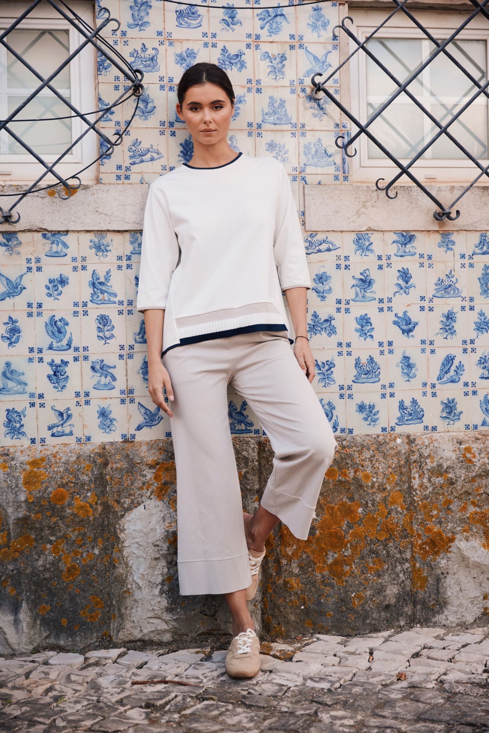 A woman stands outdoors against a blue and white tiled wall, wearing a white long-sleeve top with dark trim, light beige cropped pants, and beige sneakers.