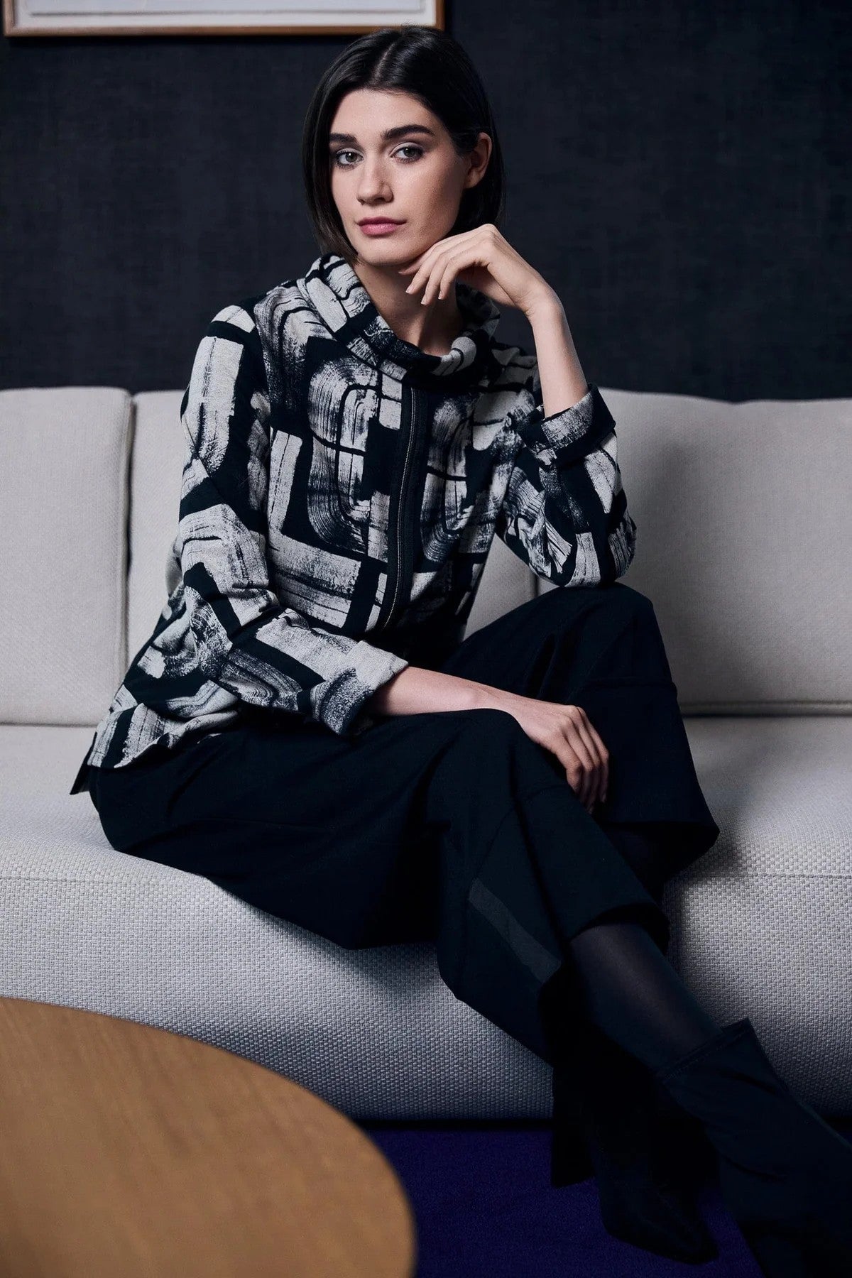 Woman sitting on a couch wearing a patterned jacket and dark pants.