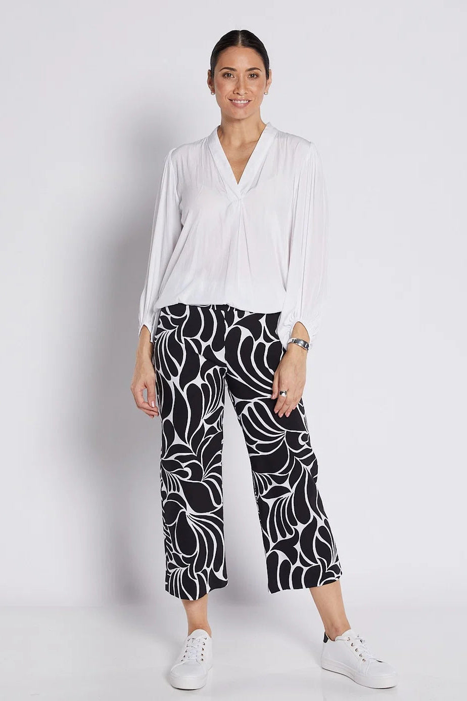 A woman modeling a white long-sleeve blouse and black cropped pants with a white abstract floral pattern, paired with white sneakers, against a light gray studio background.