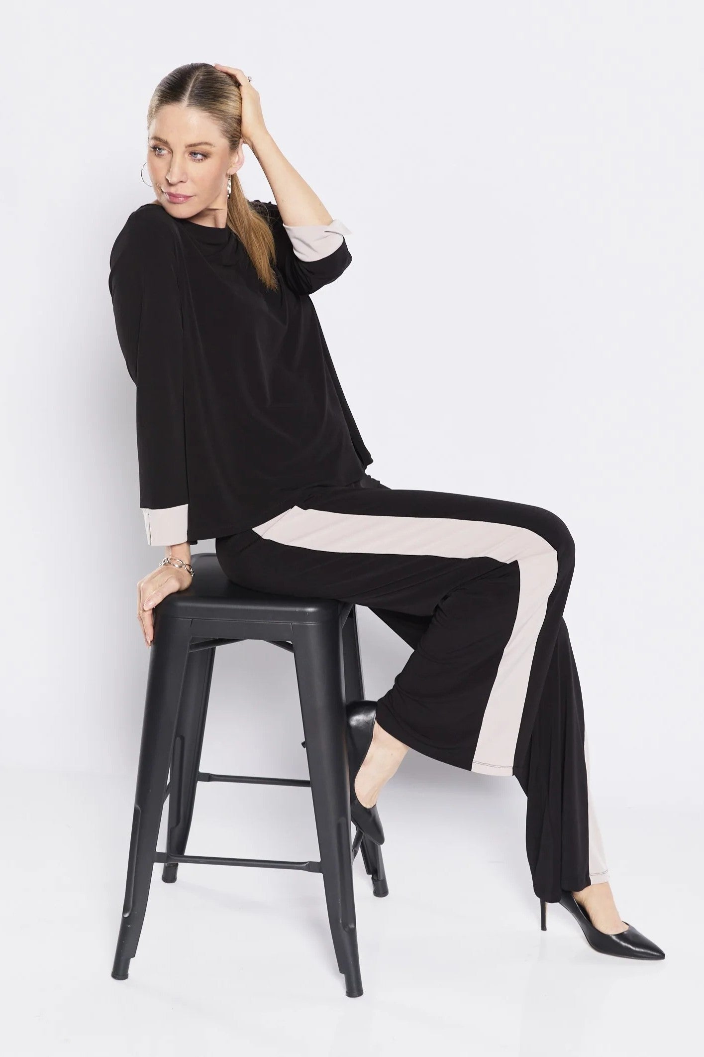 Woman sitting on a stool wearing a black outfit with white stripes against a white background
