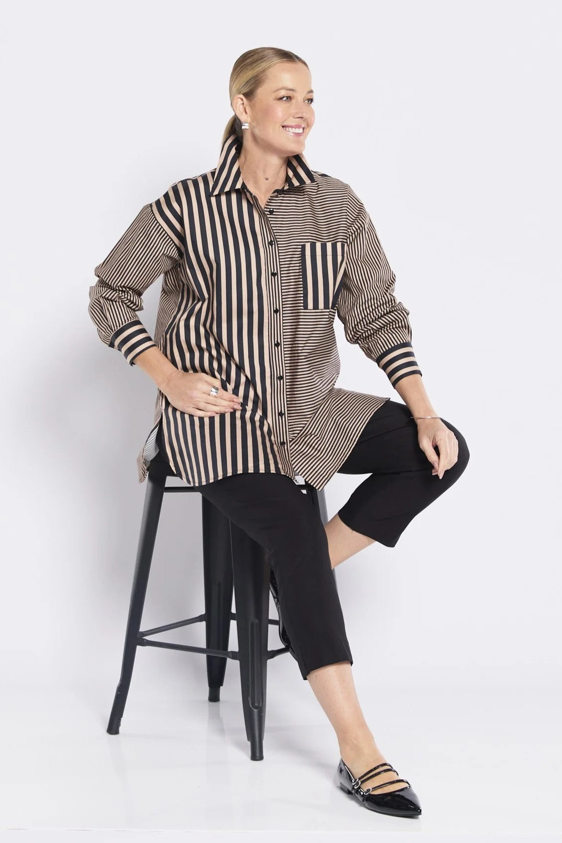 Woman wearing a patterned shirt and black pants sitting on a stool against a white background