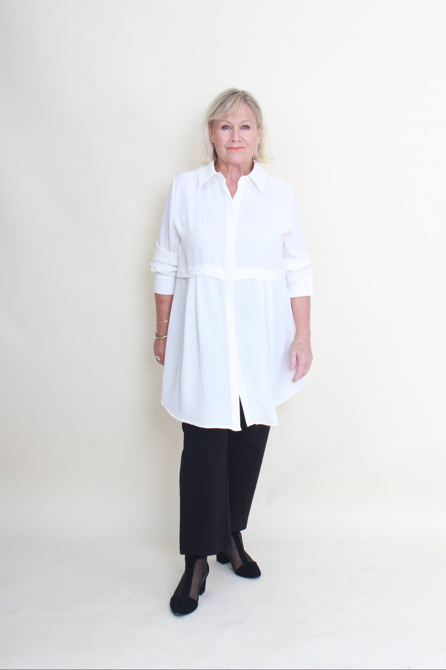 An older woman stands against a plain off-white studio background wearing a white long-sleeve button-up shirt with black wide-leg pants and black flats.