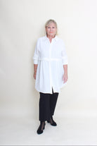An older woman stands against a plain off-white studio background wearing a white long-sleeve button-up shirt with black wide-leg pants and black flats.