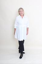 A woman modeling a white long-sleeve button-up shirt dress with a tie waist, paired with black pants and black heels, standing against a plain off-white studio background.