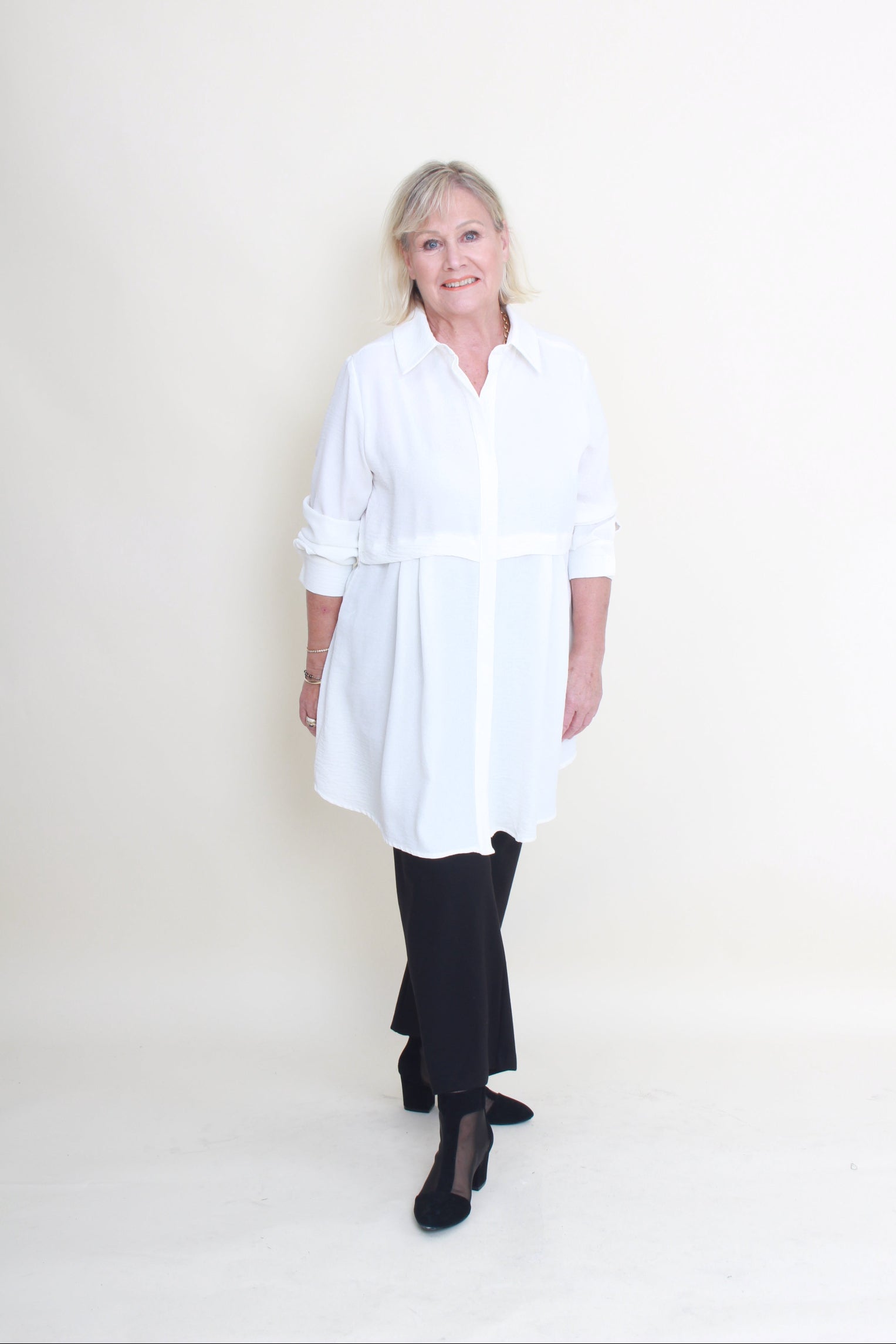 A woman modeling a white long-sleeve button-up shirt dress with a tie waist, paired with black pants and black heels, standing against a plain off-white studio background.