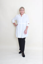 An older woman lying flat on a plain off-white studio floor, wearing a loose white long-sleeve top or dress over black pants and black shoes.