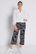 A woman modeling a white long-sleeve blouse and black cropped pants with a bold white floral pattern, paired with white sneakers, against a light gray studio background.